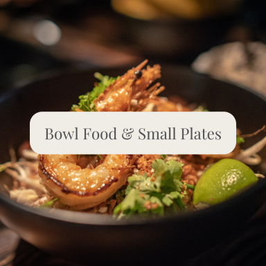 A black bowl with grilled prawns, noodles, lime, and fresh herbs.